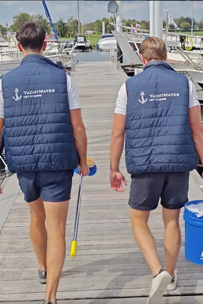 Beide eigenaren lopen over een steiger in de haven, met hun rug naar de camera. Eén houdt een schoonmaakborstel vast en de ander draagt een emmer